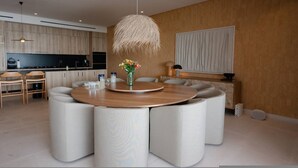 A modern dining area with a round table, white upholstered chairs, a hanging light fixture, and a kitchen in the background. - Naya 6301 Tower VI in Punta De Mita (Punta de Mita)