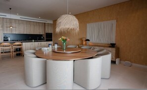 A modern dining area with a round table, white upholstered chairs, a hanging light fixture, and a kitchen in the background. - Naya 6301 Tower VI in Punta De Mita (Punta de Mita)