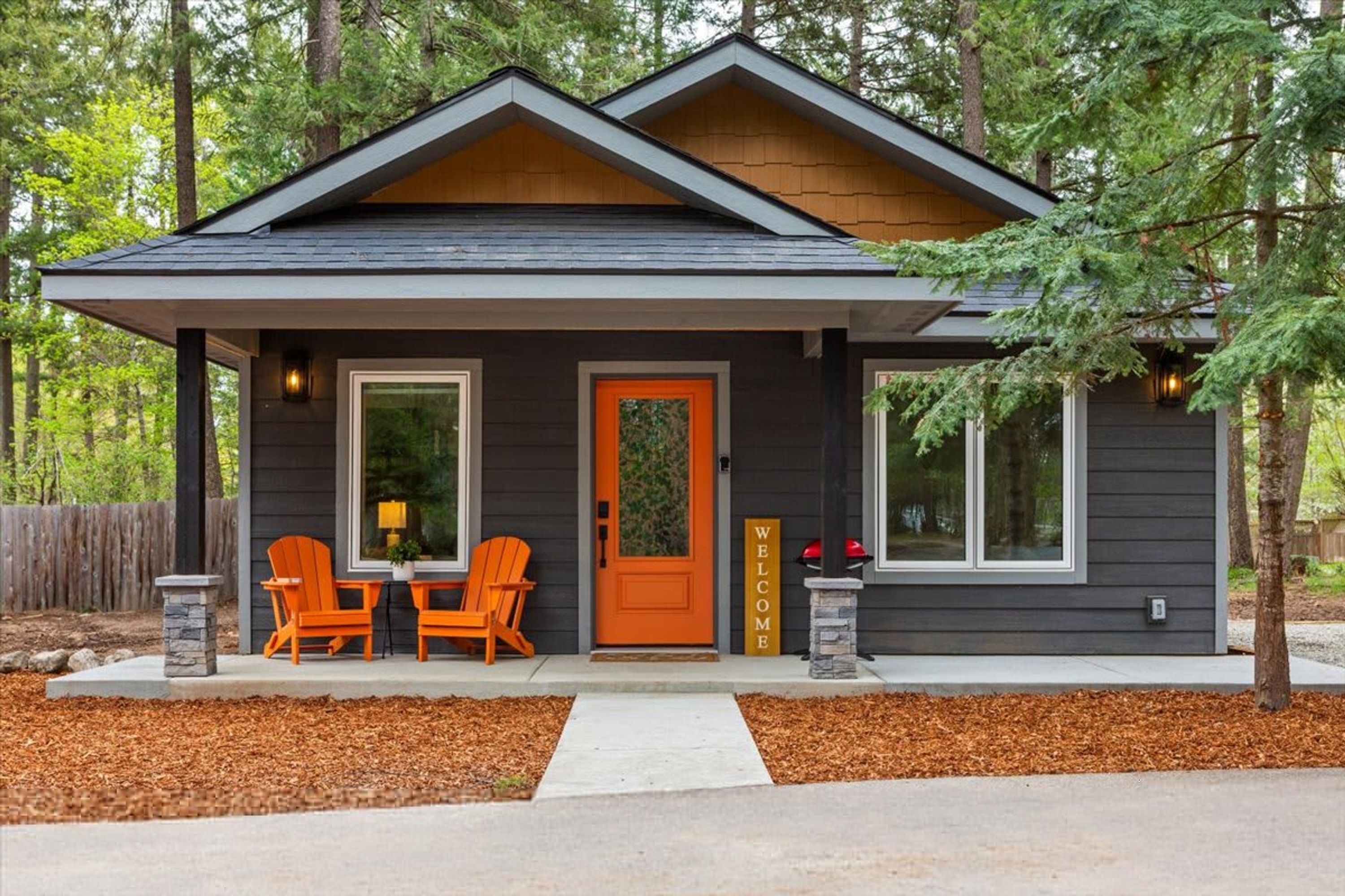 Une maison moderne à l’extérieur sombre, avec une porte orange accueillante, deux chaises sur le porche et une pancarte indiquant « BIENVENUE ».