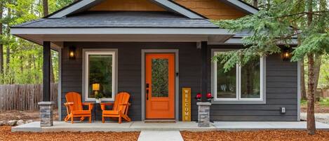 Une maison moderne à l’extérieur sombre, avec une porte orange accueillante, deux chaises sur le porche et une pancarte indiquant « BIENVENUE ».