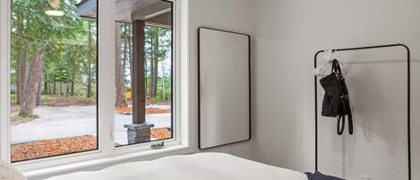 Une chambre avec une grande fenêtre, un lit, un miroir fixé au mur et un porte-manteau.