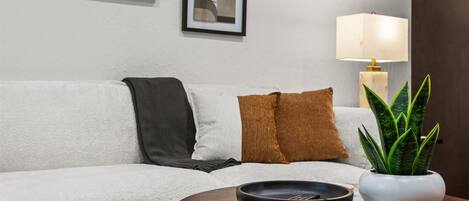 Een woonkamer met een bank, kussens, een ronde houten tafel, een zwarte schaal, een witte lamp en een potplant.
