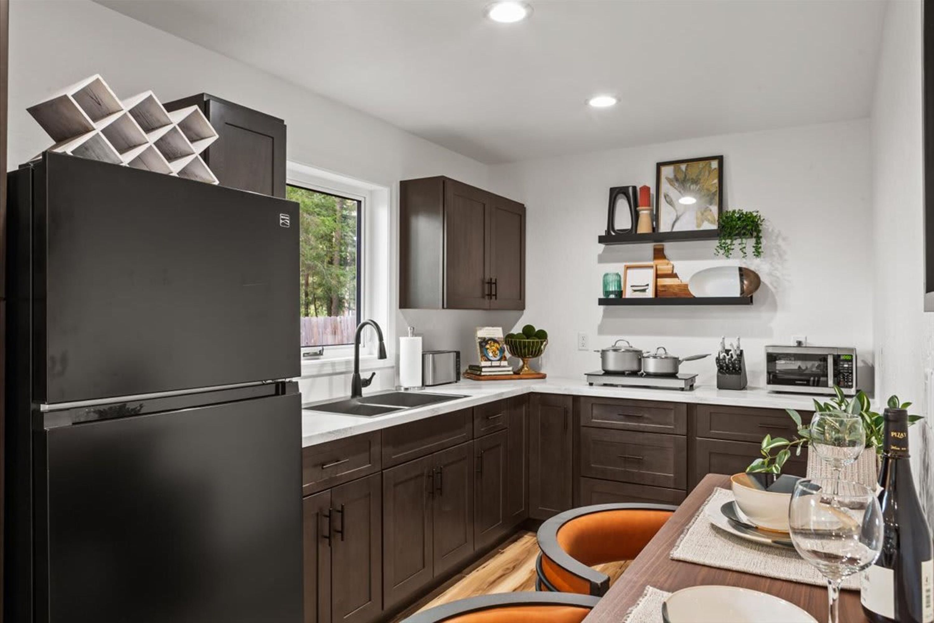 A modern kitchen with dark cabinets, a black refrigerator, a dining table with orange chairs, and a window providing a view of greenery outside.