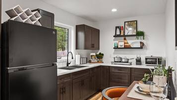 A modern kitchen with dark cabinets, a black refrigerator, a dining table with orange chairs, and a window providing a view of greenery outside.
