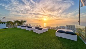 Eine Dachterrasse mit weiß gepolsterten Sitzgelegenheiten und Blick auf die Stadt bei Sonnenuntergang.