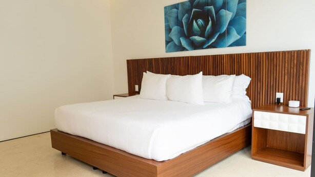 Ein modernes Hotelzimmer mit Holz-Kopfteil, weißer Bettwäsche, einem Nachttisch mit Lampe und einem an der Wand befestigten Kunstwerk.
