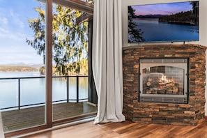 A room with a stone fireplace, a large window with a view of a lake, and a mounted TV. - Deja View - Upper Unit (Hayden)