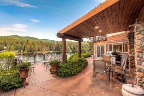 A wooden deck with outdoor seating, a stone wall, and a view of a lake and forest. - Riverfront Estate in Post Falls (Post Falls)