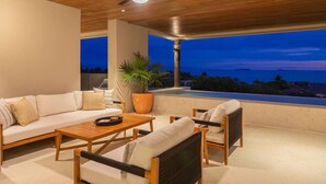 A modern outdoor living area with a sofa, chairs, a table, a ceiling fan, and a view of the ocean at dusk. - 3 bed Condo With Pool Ocean View (Punta de Mita)