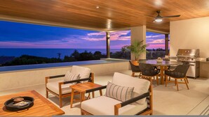 A modern outdoor seating area with a view of the ocean at sunset, wooden furniture, and a grill. - 3 bed Condo With Pool Ocean View (Punta de Mita)