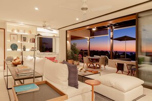 A modern living room with a sofa, coffee table, and TV. There is a balcony with outdoor furniture and a view of the sunset. - Lux 3 Bed Condo With Ocean View Pool Houskeeping (Punta de Mita)