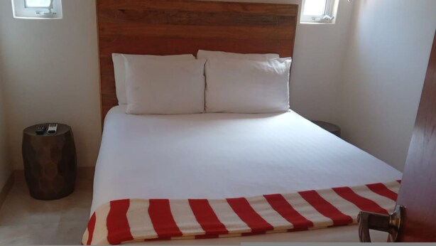 Ein Bett mit weißer Bettwäsche und einer rot-weißen gestreiften Decke, ein Holz-Kopfteil und ein kleiner Mülleimer.