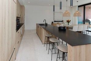 Eine moderne Küche mit Holzschränken, einer schwarzen Arbeitsplatte, Barhocker, Pendelleuchten und Blick ins Freie.