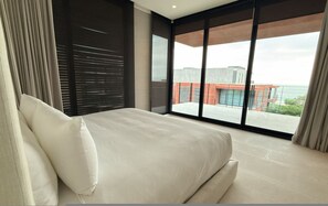 Ein modernes Schlafzimmer mit einem großen Bett, Blick auf ein Gebäude durch raumhohe Fenster und einem Balkon.