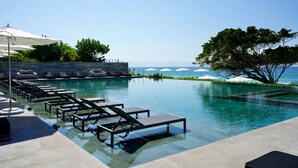 Ein Infinity-Pool mit Liegestühlen und Sonnenschirmen, ein freier Blick auf den Ozean und eine von Bäumen gesäumte Grünfläche.