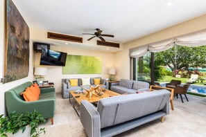 Ein modernes Wohnzimmer mit Flachbildfernseher, Deckenventilatoren und einem großen Fenster mit Blick auf einen Garten.