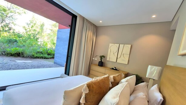A modern bedroom with a large bed, a bedside table, a lamp, and a view of greenery outside. - Naya 4101 Tower IV in Punta De Mita (Punta de Mita)