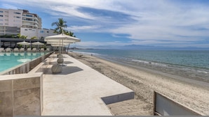 Un espace piscine aménagé au bord de la plage, avec des chaises longues et des parasols, une plage de sable, et une vue sur l’océan et les montagnes lointaines.