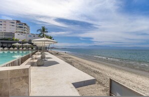 A beachside pool area with lounge chairs and umbrellas, a sandy beach, and a view of the ocean and distant mountains. - 4-bed Beachfront Lux Apartment (Bucerías)