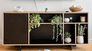 Ein modernes Wohnzimmer mit einem großen Flachbildfernseher an der Wand, einem Holztischchen mit Pflanzen und einem weißen, skulpturalen Objekt auf dem Boden.