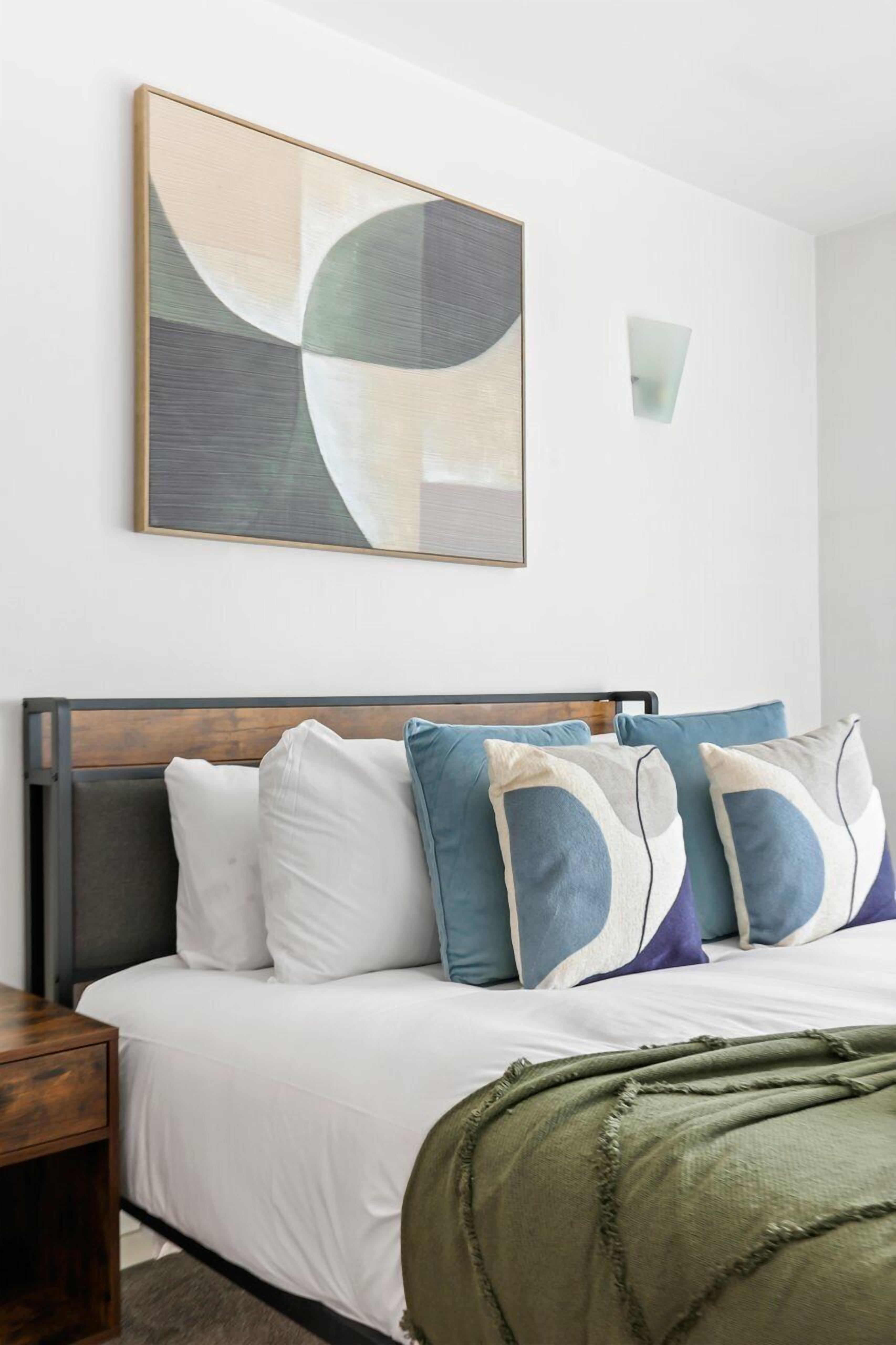 A neatly made bed with a wooden headboard, a green throw blanket, and a wall-mounted abstract painting above.