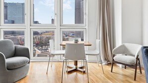Ein modernes Wohnzimmer mit Esstisch, zwei Stühlen, einer Couch und großen Fenstern mit Blick auf die Stadt.
