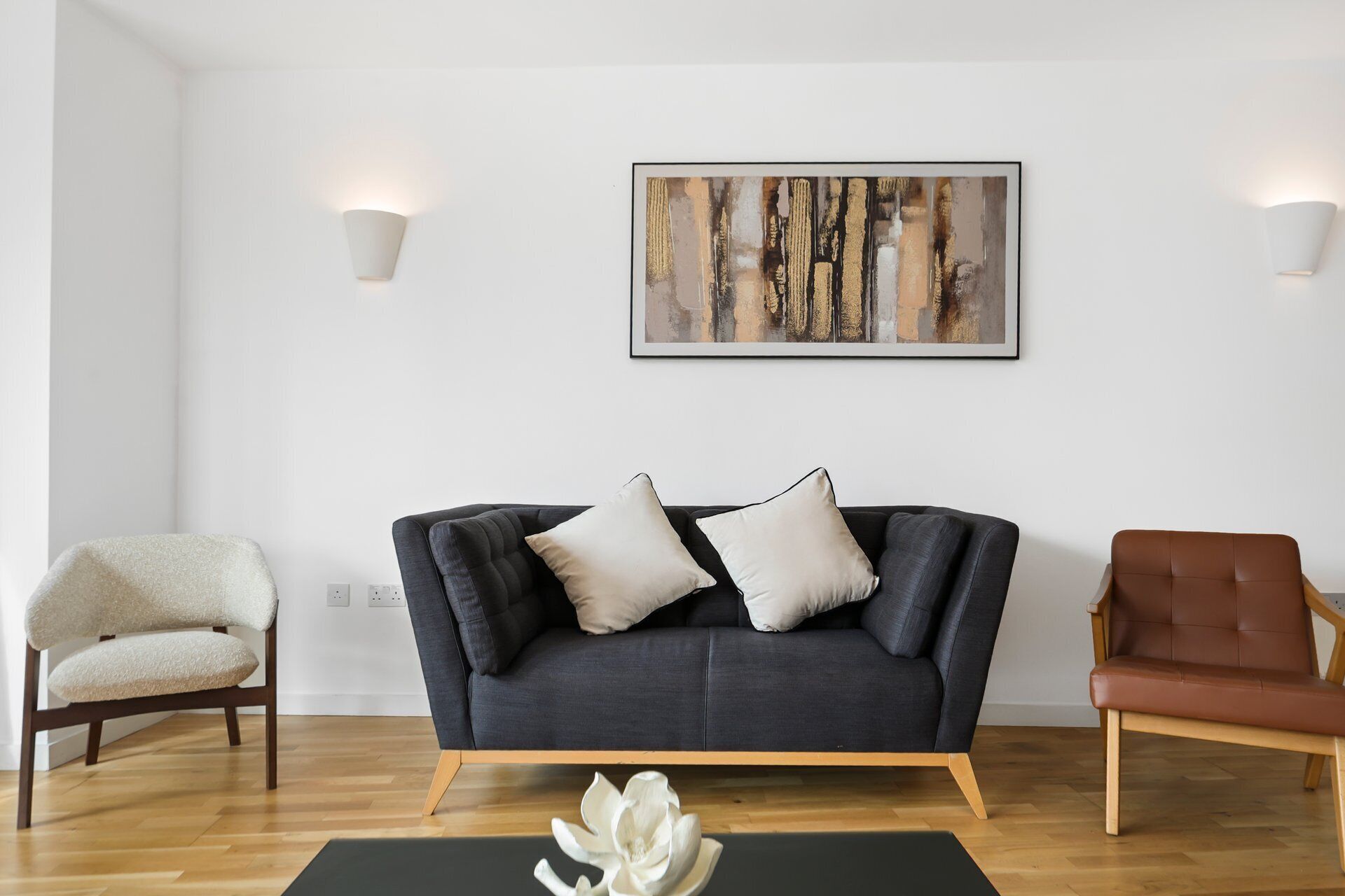 Un soggiorno con un divano grigio scuro, due cuscini bianchi, una poltrona in pelle marrone e un quadro astratto incorniciato appeso al muro.