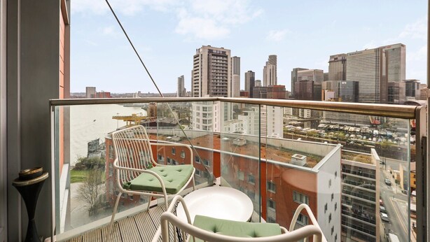Ein Balkon mit Glasgeländer, ein weißer Stuhl mit grüner Sitzauflage und Blick auf eine Skyline mit hohen Gebäuden.
