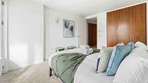 Ein Schlafzimmer mit einem Bett, einem Holzschrank, einem kleinen Hocker, einem Gemälde und einer Tür zu einem anderen Raum.
