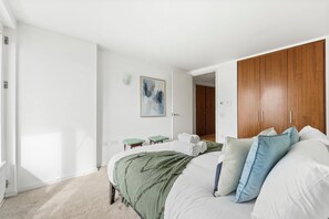 Ein Schlafzimmer mit einem Bett, einem Holzschrank, einem kleinen Hocker, einem Gemälde und einer Tür zu einem anderen Raum.