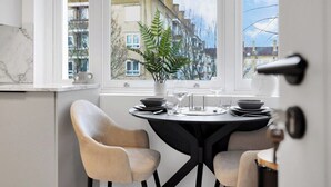 Un coin repas avec une table dressée pour deux personnes, une plante en pot et une vue sur les bâtiments par la fenêtre.