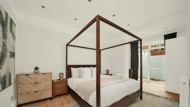 A four-poster bed with a wooden frame, a nightstand with a lamp, a dresser, and a view of the outdoors through a glass door. - Large Stylish 2 Bed Flat near Kensington (London)