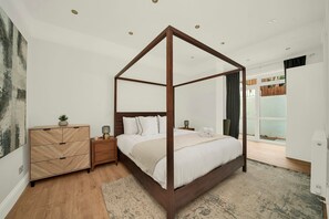 A four-poster bed with a wooden frame, a nightstand with a lamp, a dresser, and a view of the outdoors through a glass door. - Large Stylish 2 Bed Flat near Kensington (London)