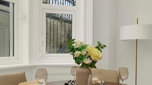 A dining table set with plates, glasses, and cutlery, a vase with flowers, and a window with a view of a fence. - Large Stylish 2 Bed Flat near Kensington (London)
