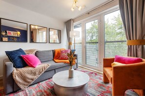 A living room with a sofa, armchair, round coffee table, and a vase with dried flowers. There are two wall-mounted mirrors, a floor lamp, and a large window with curtains. - Greenwich Station 1 min Walk Big Ben 12 min (London)