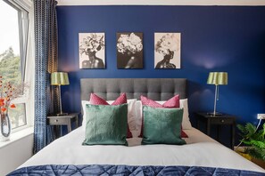 A bedroom with a bed, pillows, a headboard, wall art, a nightstand, a lamp, and a window with curtains. - Greenwich Station 1 min Walk Big Ben 12 min (London)