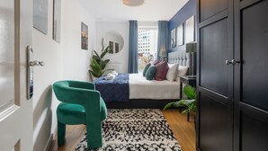 Een slaapkamer met een bed, een groene stoel, een zwart-wit vloerkleed, een raam met gordijnen en uitzicht op gebouwen.