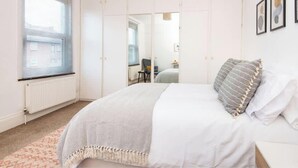 Ein Schlafzimmer mit Bett, Nachttisch, Lampe, Heizkörper, einem Fenster mit Jalousien und einer Tür zu einem anderen Raum.