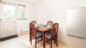 Ein Essbereich mit einem runden Tisch, gedeckt für vier Personen, Holzsesseln mit gestreiften Kissen, einem weißen Kühlschrank und einem Fenster mit Jalousien.