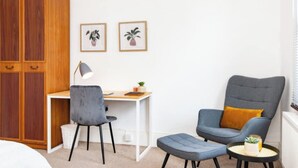 Ein Zimmer mit Schreibtisch, zwei Stühlen, einem Kleiderschrank und zwei gerahmten Bildern an der Wand.