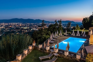 A pool with lounge chairs and umbrellas, surrounded by greenery and overlooking a cityscape at dusk. - Superior Garden Borgo di Matraia (Capannori)