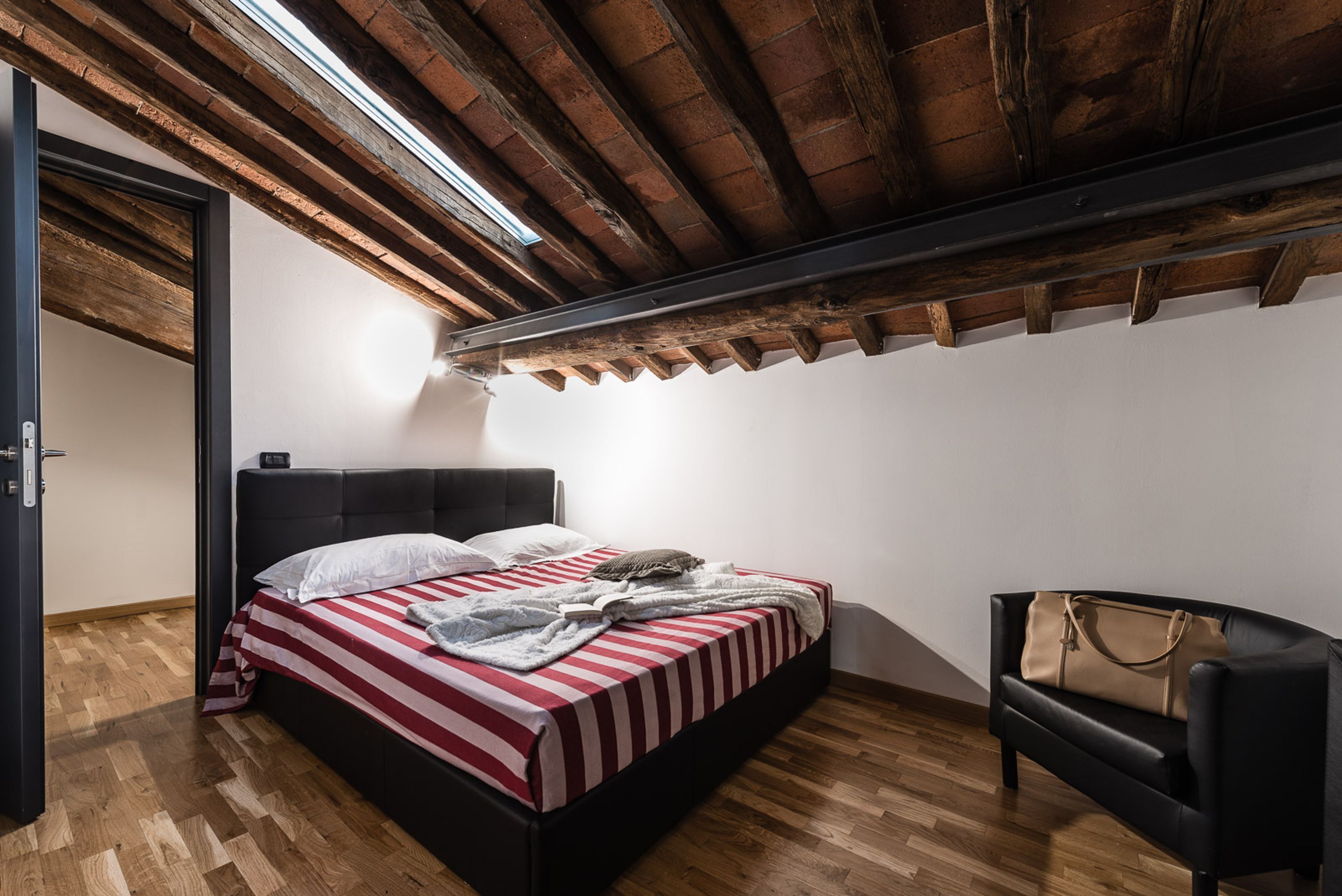 A bedroom with a bed, a chair, a wooden floor, a ceiling with exposed beams, and a door.