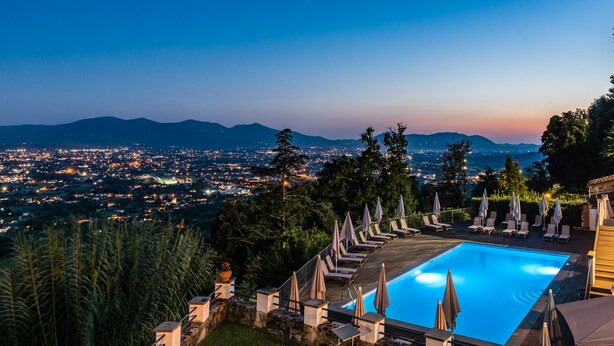 Ein Pool mit Liegestühlen und Sonnenschirmen, umgeben von Grün und mit Blick auf eine Stadtlandschaft in der Abenddämmerung.