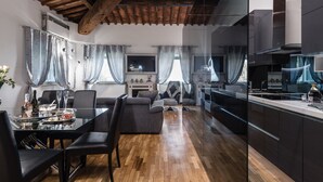 Eine moderne Küche mit dunklen Schränken und einer Holzbalkendecke, ein Essbereich mit einem Glastisch und Stühlen sowie ein Wohnzimmer mit einer Couch und Vorhängen.