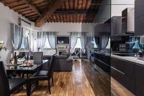 A modern kitchen with dark cabinets and a wooden ceiling, a dining area with a glass table and chairs, and a living room with a sofa and curtains. - Giotto Luxury Apartment Villa Guinigi (Capannori)