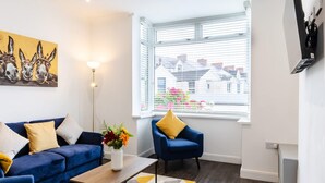 A living room with a blue sofa, a coffee table, a blue armchair, a painting, and a window with blinds. - 1-min Walk to High St 3-min to Mumbles Beach (Swansea)