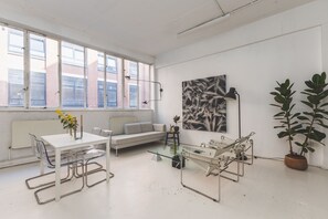 A modern room with a glass table, metal chairs, a sofa, a radiator, and large windows. - Modern Flat in the Heart of Bethnal Green (London)