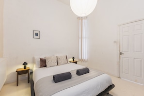 A bedroom with a bed, two bedside tables, a nightstand, a lamp, a picture on the wall, and a door. - Modern Flat in the Heart of Bethnal Green (London)