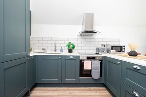 A modern kitchen with teal cabinets, white tiles, a stainless steel range hood, a microwave, and a dishwasher. - Omega Acton Top Floor (London)