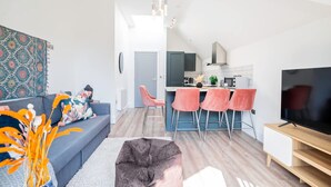 Een moderne woonkamer met een bank, poef en een televisie. Er is een keukenhoek met een eettafel en stoelen.